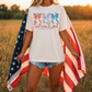 A woman stands in a field, holding an American flag and wearing a white t-shirt with a graphic design.