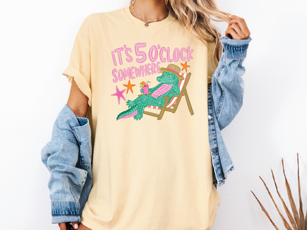 A person wearing a yellow t-shirt with a graphic of a green alligator sitting in a chair and the text "IT'S 50 CLOCK SOMEWHERE" printed on it.