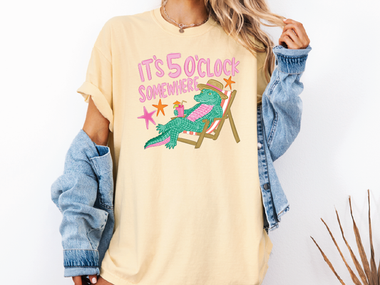 A person wearing a yellow t-shirt with a graphic of a green alligator sitting in a chair and the text "IT'S 50 CLOCK SOMEWHERE" printed on it.