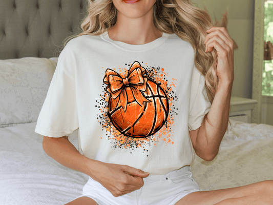 A woman wearing a white t-shirt with an orange basketball graphic and a bow on it, sitting on a bed.