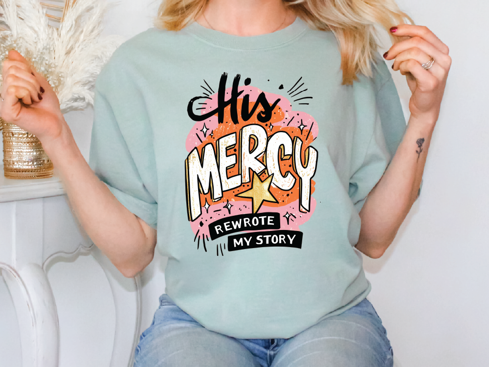 A woman wearing a light blue t-shirt with a graphic design that says "His Mercy" and "Rewrote My Story".