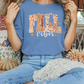 A woman wearing a blue t-shirt with the text "FALL VIBES" is sitting on a bench surrounded by fall decorations, including pumpkins and a "Home is where the heart is" sign.