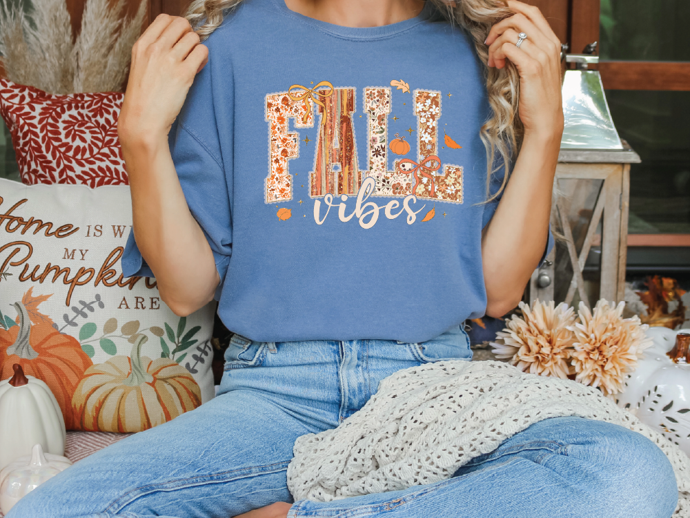 A woman wearing a blue t-shirt with the text "FALL VIBES" is sitting on a bench surrounded by fall decorations, including pumpkins and a "Home is where the heart is" sign.