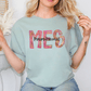 A woman wearing a light blue t-shirt with the text "MES" printed on it, posing with her hands behind her back.