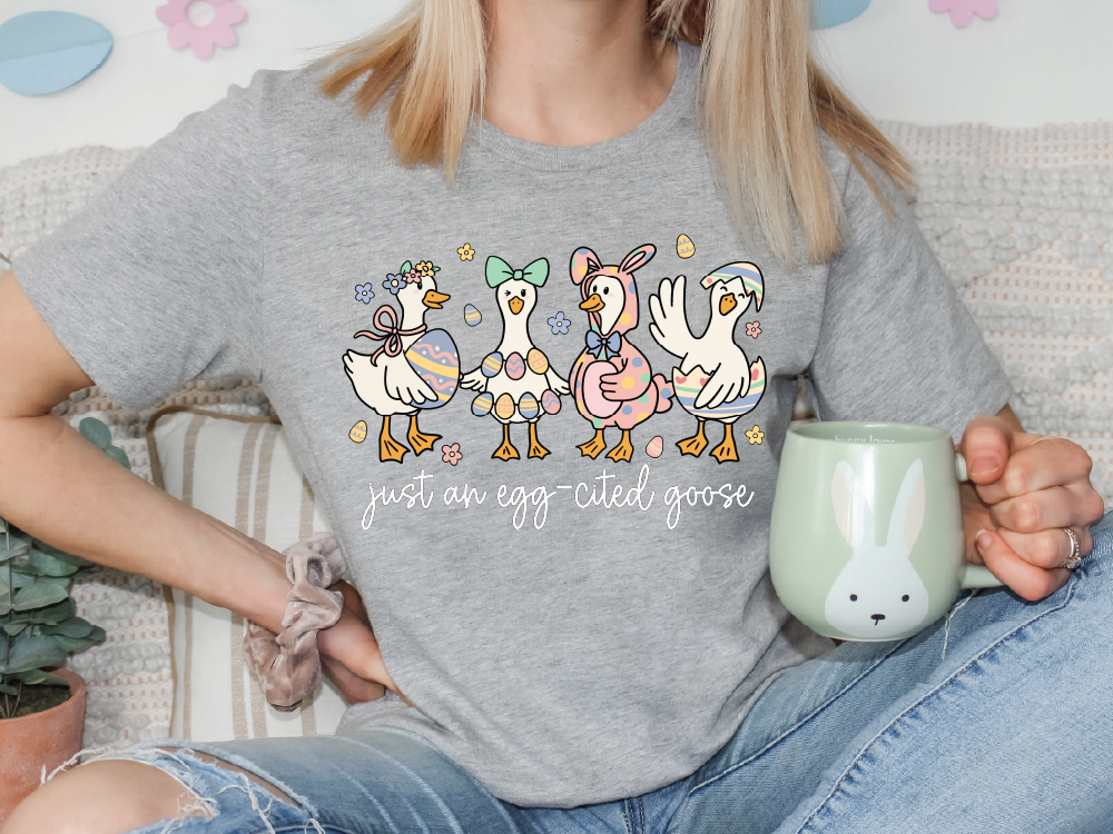 A woman wearing a gray t-shirt with a graphic of four cartoon ducks and the text "Just an egg cited goose" is sitting on a couch, holding a green mug.