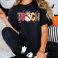 A person wearing a black t-shirt with a graphic design of a ghost and the word "Teach" on it, sitting on a couch with Halloween decorations in the background.