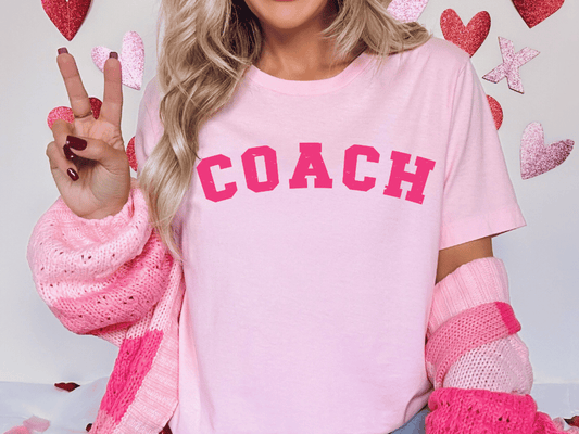 A woman wearing a pink t-shirt with the word "COACH" printed on it, making a peace sign with her hand. She is also wearing a pink sweater and has her hair styled in loose waves.