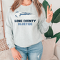 A woman wearing a gray sweatshirt with the text "LONG COUNTY BLUETIDE" printed on it, standing in front of a plant.