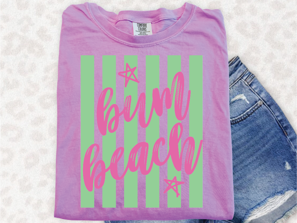 A pink t-shirt with the text "Bum Beach" printed on it, along with green and white striped patterns, is displayed alongside a pair of blue denim shorts.
