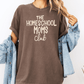 A person wearing a brown t-shirt with the text "The Homeschool Moms Club" printed on it, along with a denim jacket and a plant in the background.