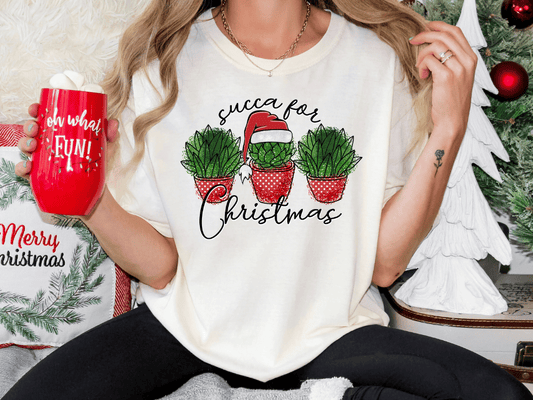 A person wearing a white t-shirt with a graphic design of three potted succulents and a Santa hat, holding a red mug with the text "oh what fun!"