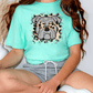 A person wearing a turquoise t-shirt with a graphic of a bulldog and a leopard print design, sitting on the floor with their legs crossed.