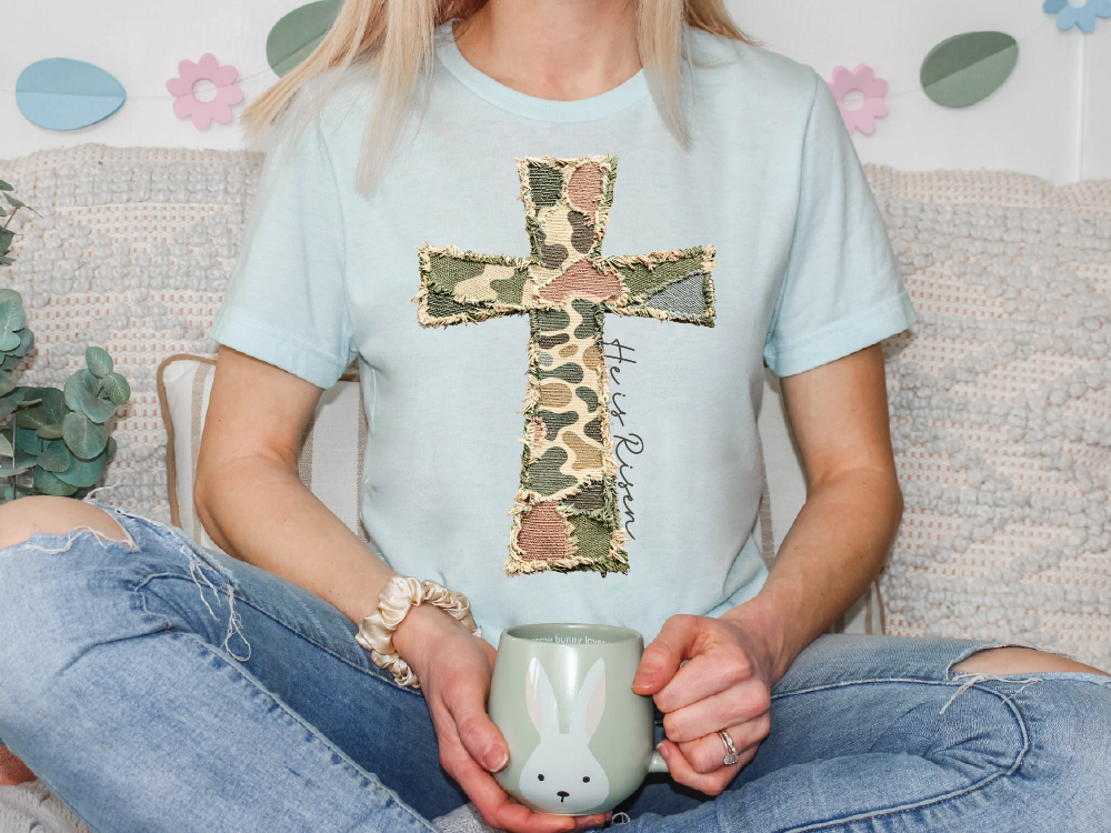 A person is sitting on a couch, wearing a light blue t-shirt with a camouflage cross design, and holding a mug with a bunny face.