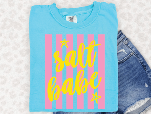 A blue t-shirt with a yellow and pink striped design and the text "salt babe" printed on it, along with a pair of denim shorts.