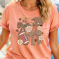 A woman wearing a peach-colored t-shirt with a graphic design featuring a football, flowers, and a bow.