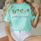 A woman wearing a mint green t-shirt with a graphic of potted plants and the text "just one make" is sitting on a bed.