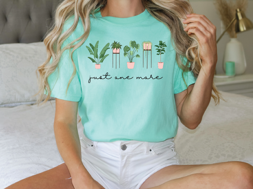 A woman wearing a mint green t-shirt with a graphic of potted plants and the text "just one make" is sitting on a bed.