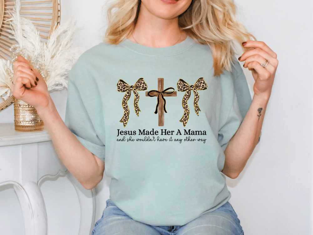 A woman wearing a light blue t-shirt with a graphic design of leopard print bows and text that reads "Jesus Made Her A Mama and she wouldn't have it any other way".