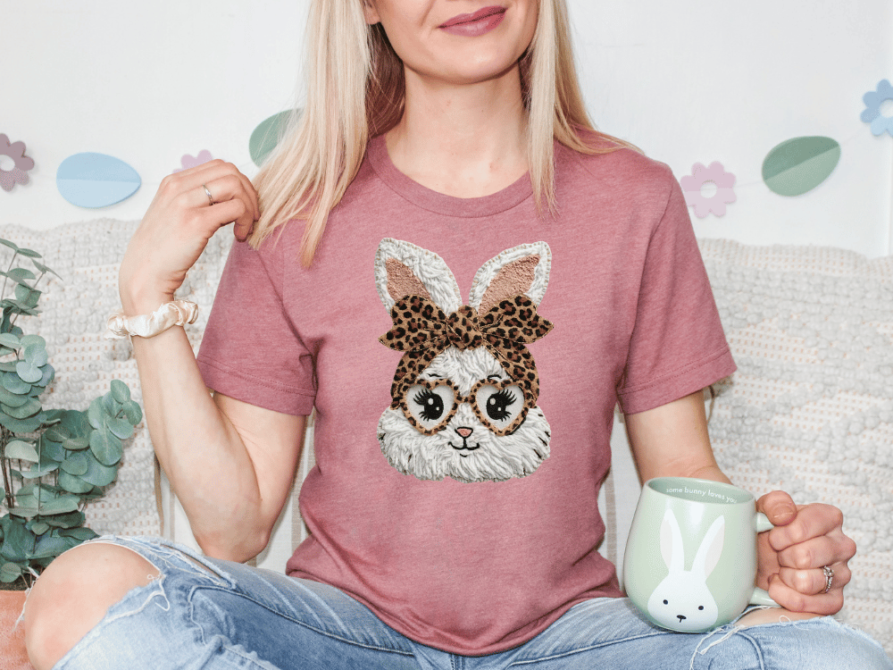 A woman wearing a pink t-shirt with a bunny design and glasses is sitting on a couch, holding a mug.