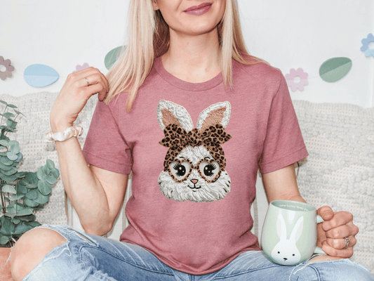A woman wearing a pink t-shirt with a bunny design and glasses is sitting on a couch, holding a mug.
