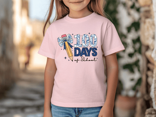 A young girl wearing a pink t-shirt with a graphic design that says "100 DAYS of School" and features a pencil and a bow.