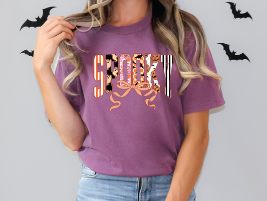 A woman wearing a purple t-shirt with the word "SPOOKY" printed on it, standing in front of a wall with bats.