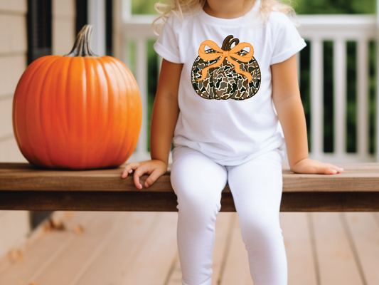 A young girl wearing a white t-shirt with a camouflage pumpkin design sits on a wooden bench next to a large orange pumpkin.