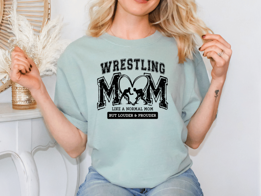 A woman wearing a light blue t-shirt with a graphic design that says "Wrestling Mom like a normal mom but louder & proud".