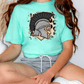 A person wearing a turquoise t-shirt with a graphic of a helmet and a leopard print design, sitting on the floor with their legs crossed.