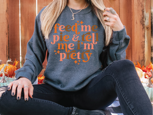 a woman sitting on the ground wearing a sweatshirt that says feed me pie and tell